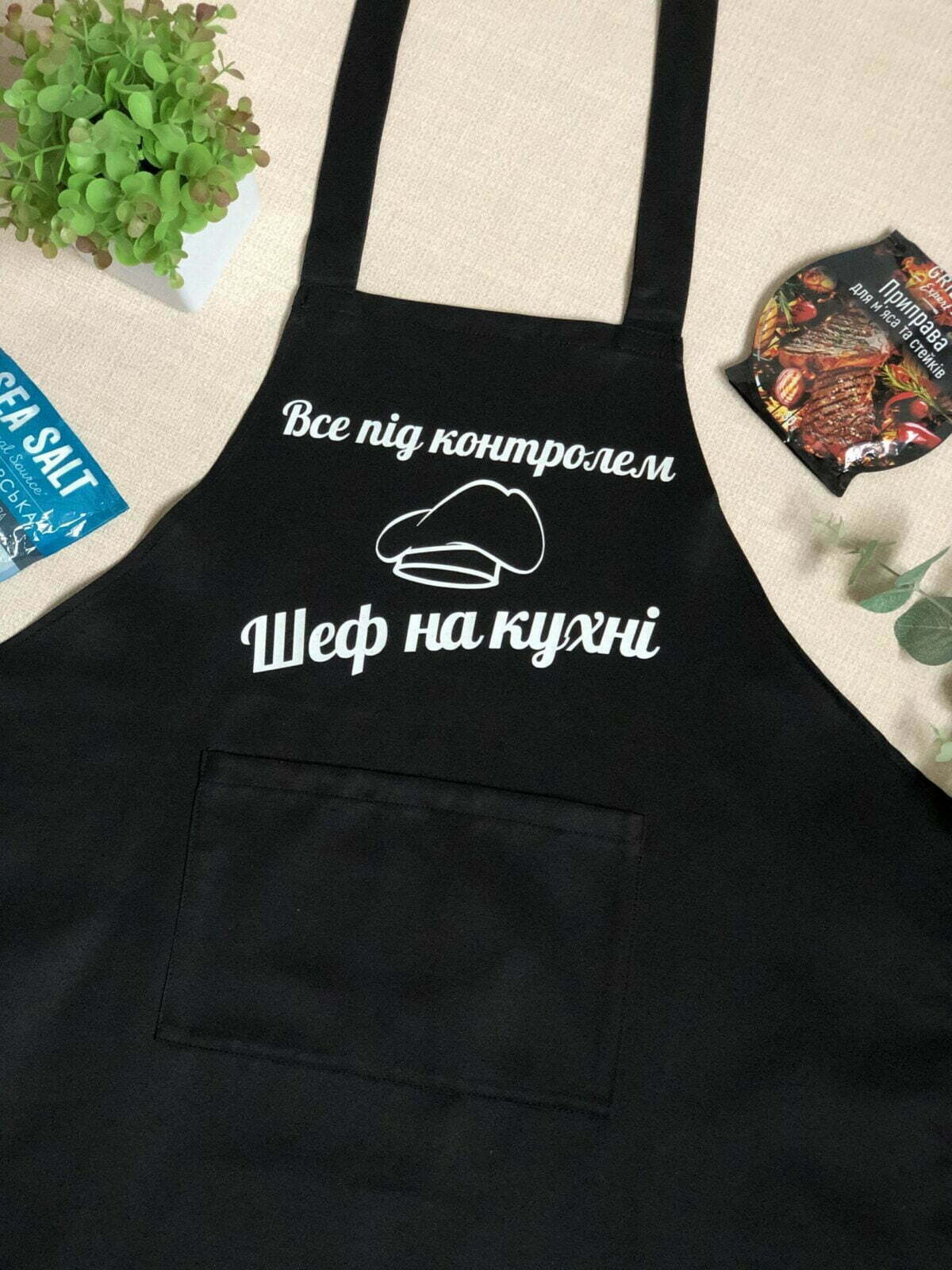 На даному зображенні представлений фартух з веселим принтом "Шеф на кухні". Фартух має чорний колір і виготовлений з якісного матеріалу. На фартушку зображено кухонні прилади, зелень та напис "Шеф на кухні". Фартух має регульовані зав'язки на шиї та поясі, що дозволяє налаштувати його під потрібний розмір. Цей фартух стане відмінним подарунком для тих, хто любить готувати та прикрашати свій дім.