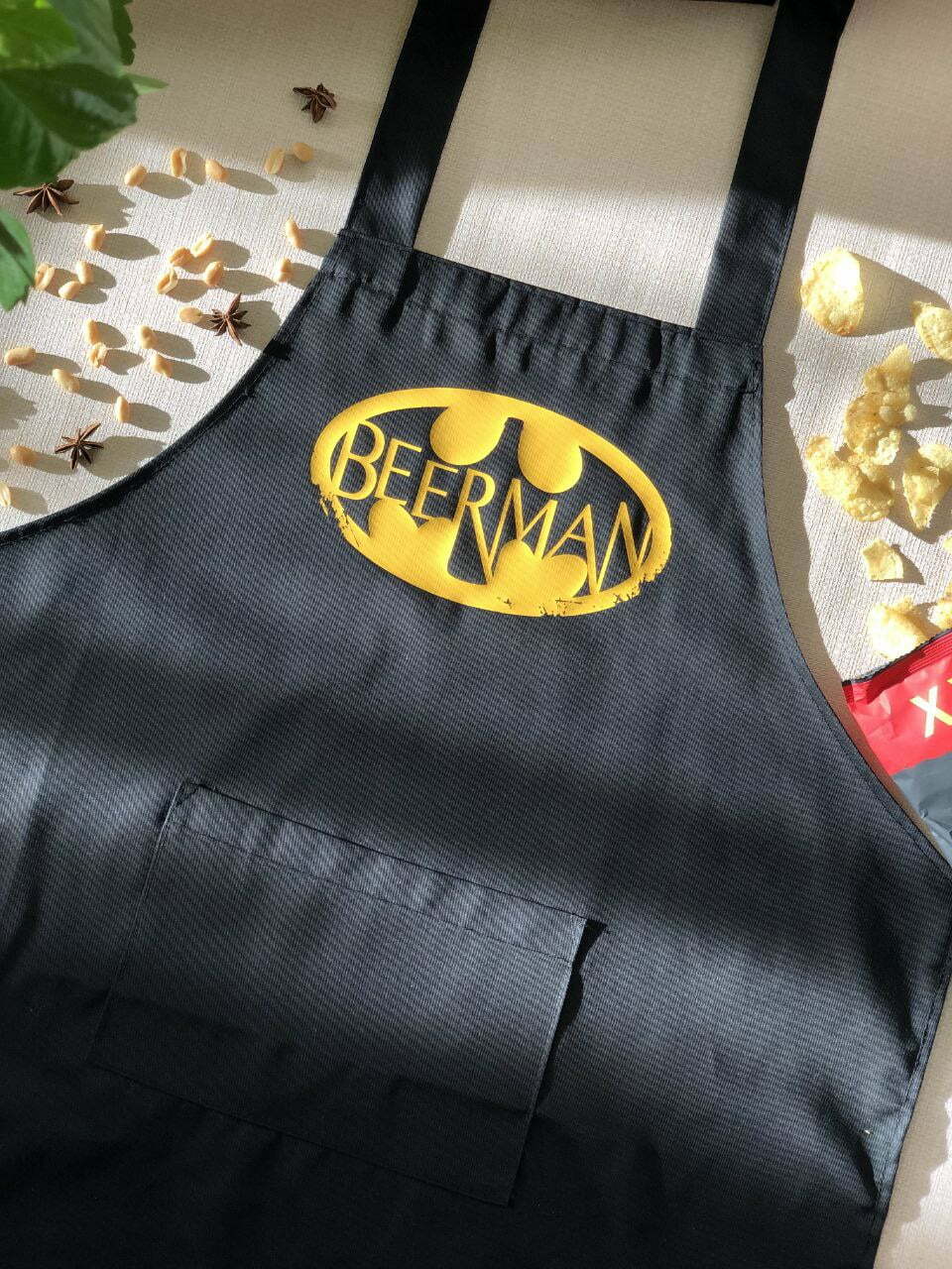 На даному зображенні представлений фартух з веселим принтом "Beerman". Фартух має чорний колір і виготовлений з якісного матеріалу. На фартушку зображено зелень та напис "Beerman". Фартух має регульовані зав'язки на шиї та поясі, що дозволяє налаштувати його під потрібний розмір. Цей фартух стане відмінним подарунком для тих, хто любить готувати та прикрашати свій дім.
