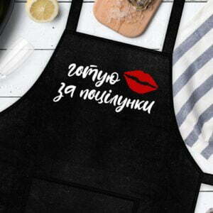 На даному зображенні представлений фартух з веселим принтом "Готую за поцілунки". Фартух має чорний колір і виготовлений з якісного матеріалу. На фартушку зображено кухонні прилади, зелень та напис "Готую за поцілунки". Фартух має регульовані зав'язки на шиї та поясі, що дозволяє налаштувати його під потрібний розмір. Цей фартух стане відмінним подарунком для тих, хто любить готувати та прикрашати свій дім.