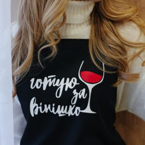 На даному зображенні представлений фартух з веселим принтом "Готую за вінішко". Фартух має чорний колір і виготовлений з якісного матеріалу. На фартушку зображено кухонні прилади, зелень та напис "Готую за вінішко". Фартух має регульовані зав'язки на шиї та поясі, що дозволяє налаштувати його під потрібний розмір. Цей фартух стане відмінним подарунком для тих, хто любить готувати та прикрашати свій дім.