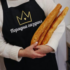 На даному зображенні представлений фартух з веселим принтом "Порядна газдиня". Фартух має чорний колір і виготовлений з якісного матеріалу. На фартушку зображено кухонні прилади, зелень та напис "Порядна газдиня". Фартух має регульовані зав'язки на шиї та поясі, що дозволяє налаштувати його під потрібний розмір. Цей фартух стане відмінним подарунком для тих, хто любить готувати та прикрашати свій дім.