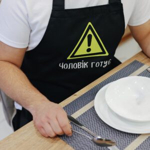 На даному зображенні представлений фартух з веселим принтом "Чоловік готує". Фартух має чорний колір і виготовлений з якісного матеріалу. На фартушку зображено кухонні прилади, зелень та напис "Чоловік готує". Фартух має регульовані зав'язки на шиї та поясі, що дозволяє налаштувати його під потрібний розмір. Цей фартух стане відмінним подарунком для тих, хто любить готувати та прикрашати свій дім.