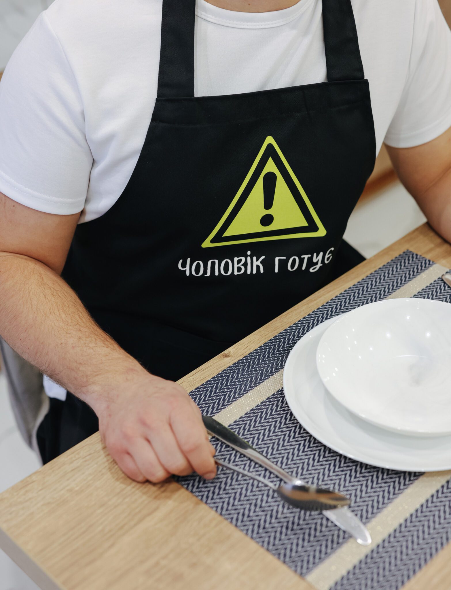 На даному зображенні представлений фартух з веселим принтом "Чоловік готує". Фартух має чорний колір і виготовлений з якісного матеріалу. На фартушку зображено кухонні прилади, зелень та напис "Чоловік готує". Фартух має регульовані зав'язки на шиї та поясі, що дозволяє налаштувати його під потрібний розмір. Цей фартух стане відмінним подарунком для тих, хто любить готувати та прикрашати свій дім.