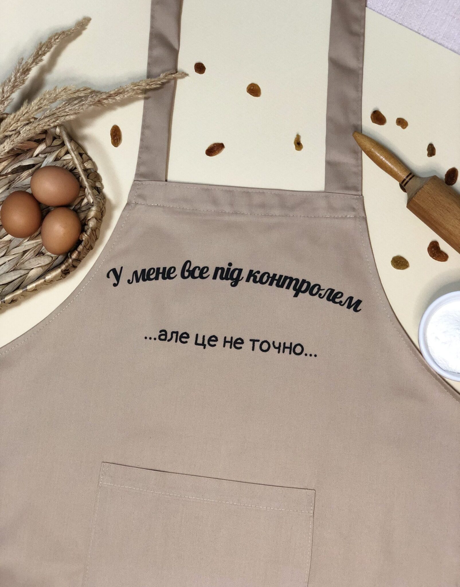 На даному зображенні представлений фартух з веселим принтом "Все під контролем". Фартух має бежевий колір і виготовлений з якісного матеріалу. На фартушку зображено кухонні прилади, зелень та напис "Все під контролем". Фартух має регульовані зав'язки на шиї та поясі, що дозволяє налаштувати його під потрібний розмір. Цей фартух стане відмінним подарунком для тих, хто любить готувати та прикрашати свій дім.