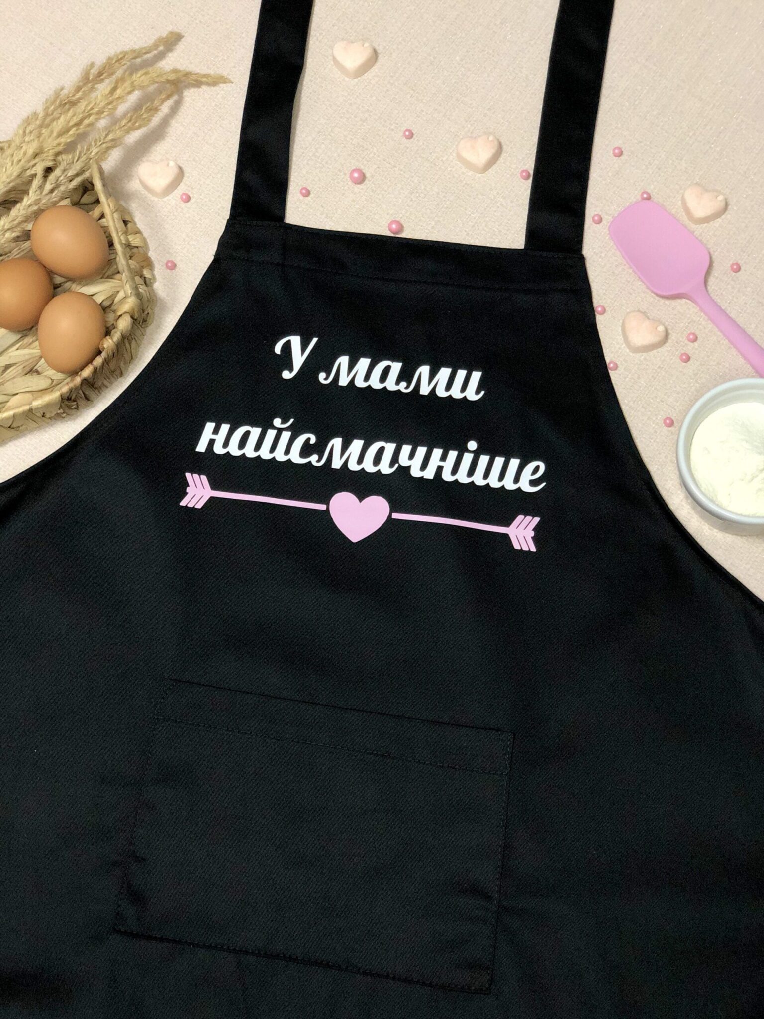 На даному зображенні представлений фартух з веселим принтом "У мами найсмачніше". Фартух має чорний колір і виготовлений з якісного матеріалу. На фартушку зображено кухонні прилади, зелень та напис "У мами найсмачніше", що матуся готує". Фартух має регульовані зав'язки на шиї та поясі, що дозволяє налаштувати його під потрібний розмір. Цей фартух стане відмінним подарунком для тих, хто любить готувати та прикрашати свій дім.