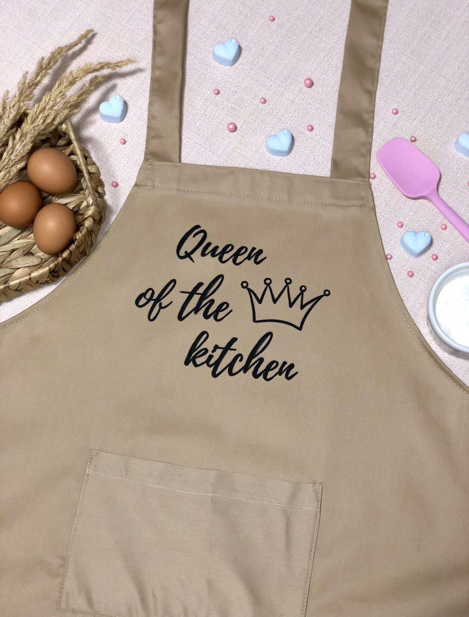 На даному зображенні представлений фартух з веселим принтом "Queen of the kitchen". Фартух має бежевий колір і виготовлений з якісного матеріалу. На фартушку зображено кухонні прилади, зелень та напис "Queen of the kitchen". Фартух має регульовані зав'язки на шиї та поясі, що дозволяє налаштувати його під потрібний розмір. Цей фартух стане відмінним подарунком для тих, хто любить готувати та прикрашати свій дім.