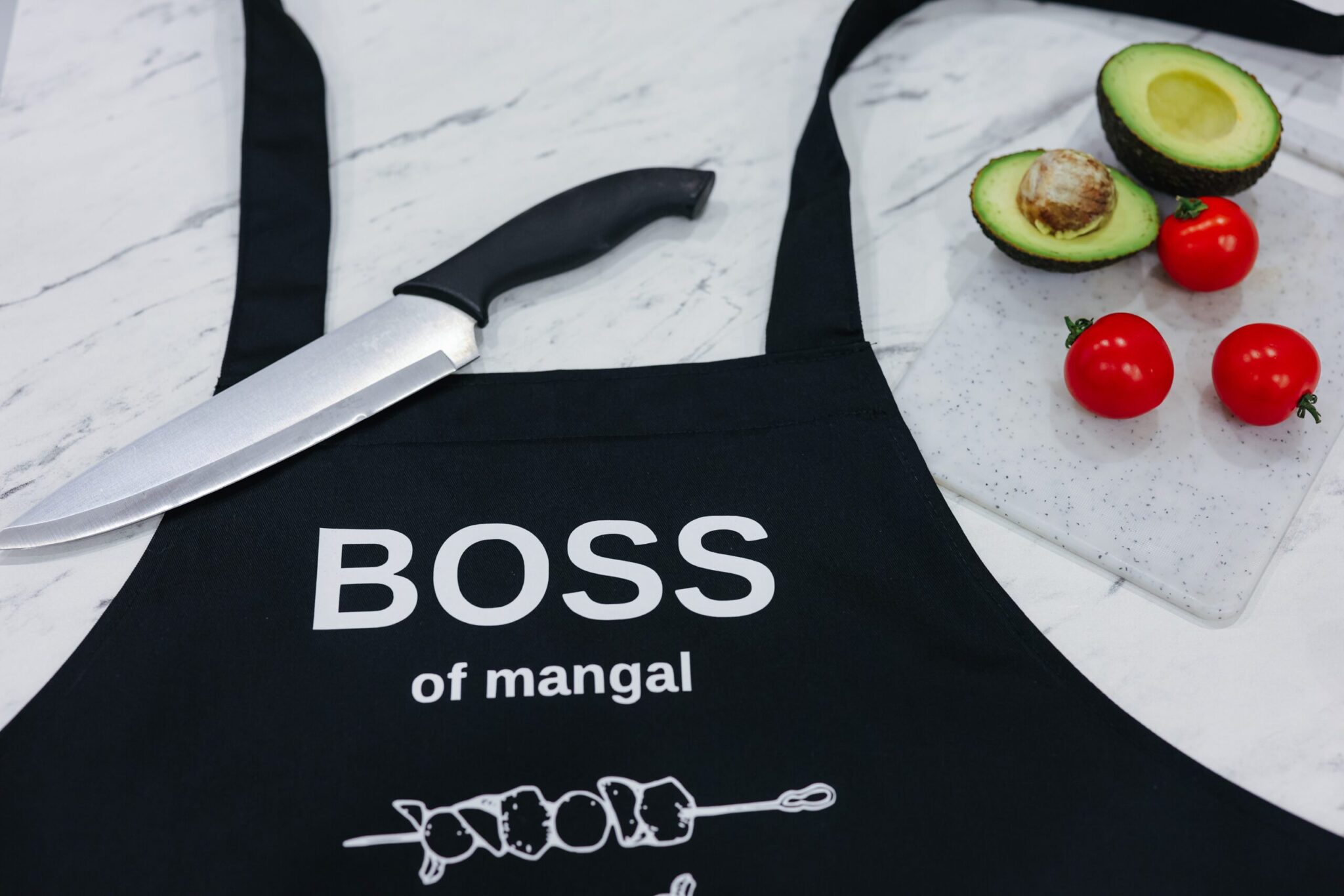 На даному зображенні представлений фартух з веселим принтом "Boss of mangal". Фартух має чорний колір і виготовлений з якісного матеріалу. На фартушку зображено кухонні прилади, зелень та напис "Boss of mangal". Фартух має регульовані зав'язки на шиї та поясі, що дозволяє налаштувати його під потрібний розмір. Цей фартух стане відмінним подарунком для тих, хто любить готувати та прикрашати свій дім.